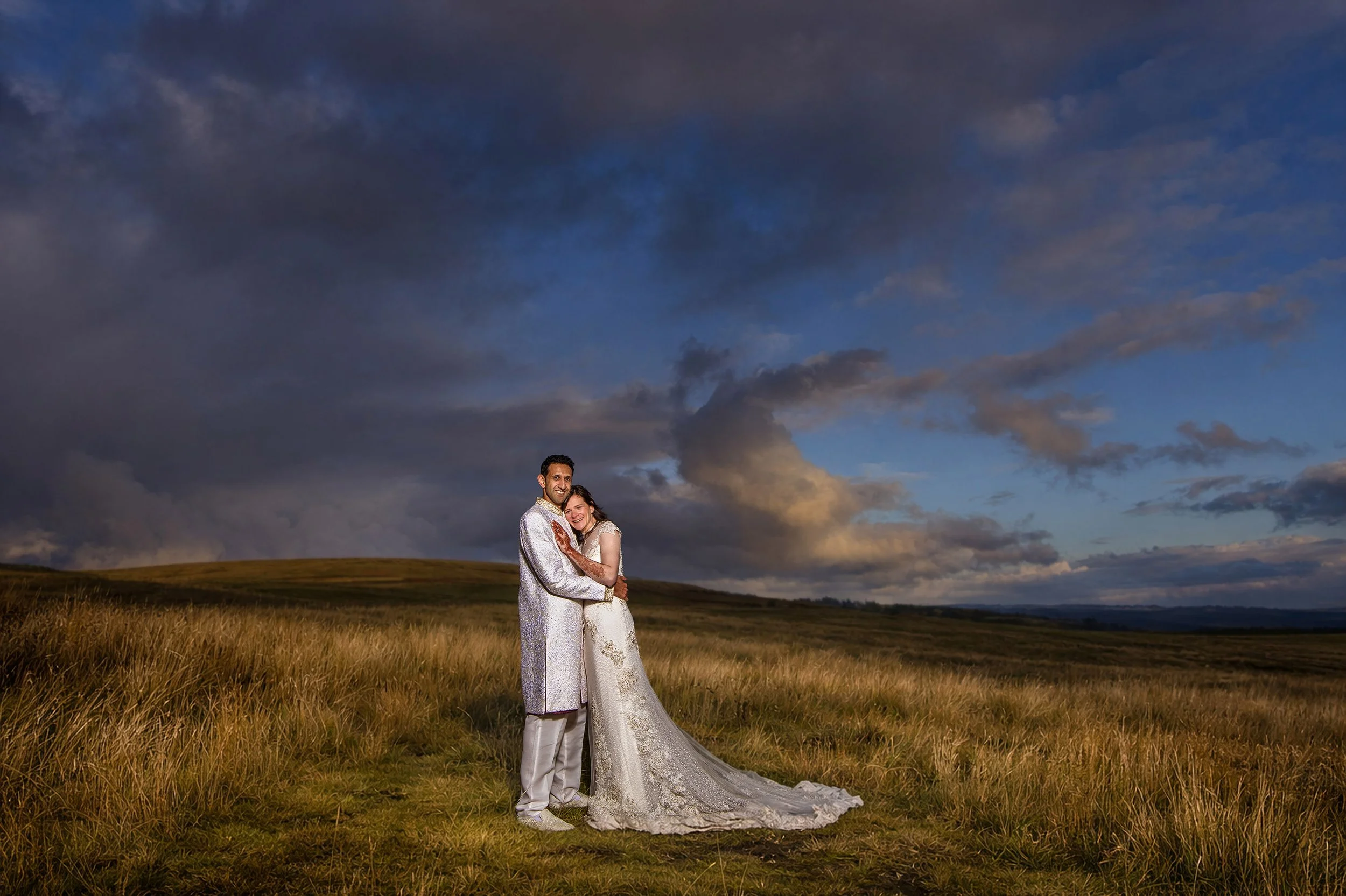 A couple in wedding attire embracing in an open grassy field at sunset with dark clouds in the sky.