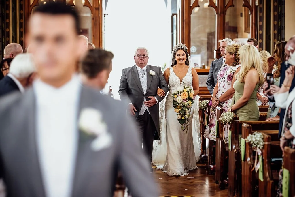 A bride walking down the aisle with her father in a church during her wedding, surrounded by guests in seats decorated with flowers.