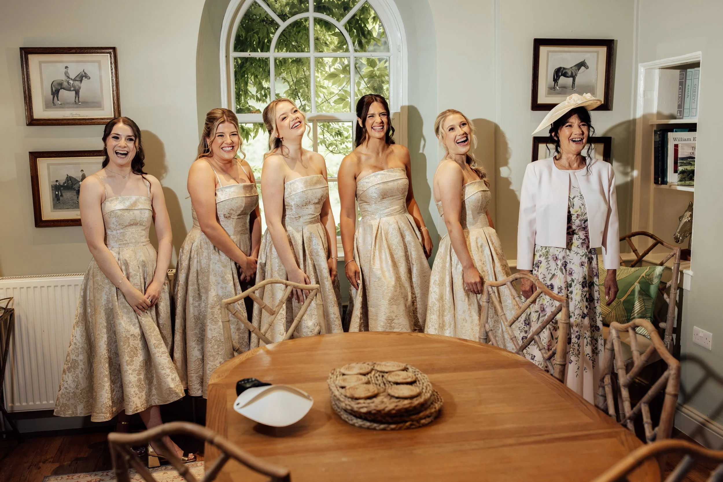 Six women, five in matching gold dresses and one in a white jacket and floral dress, are standing inside a room with cream-colored walls and framed horse pictures, laughing and smiling.