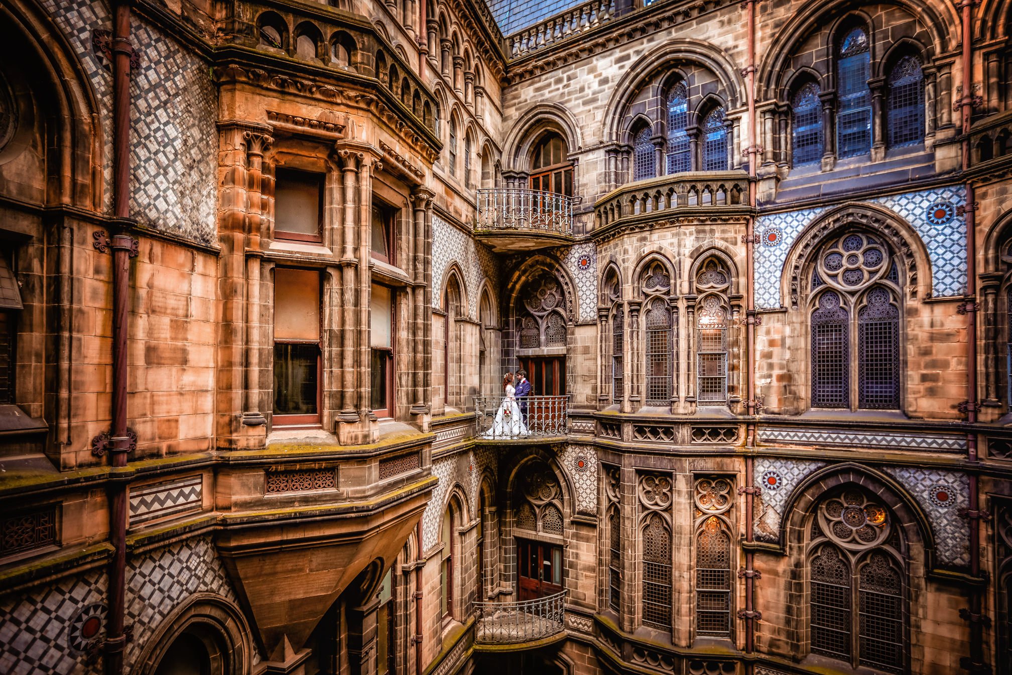 A bride and groom standing on a small balcony amidst the ornate stone walls of a historic building with Gothic-style windows and detailed architecture.