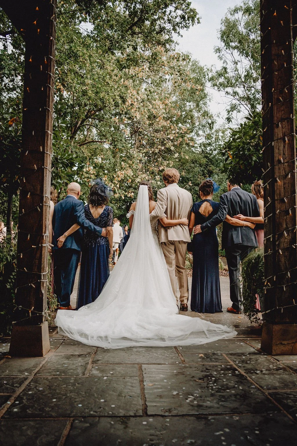 A group of wedding guests in formal attire standing outdoors, seen from behind, with a bride in a white wedding gown and veil and a groom in a light-colored suit in the center, surrounded by greenery and trees.