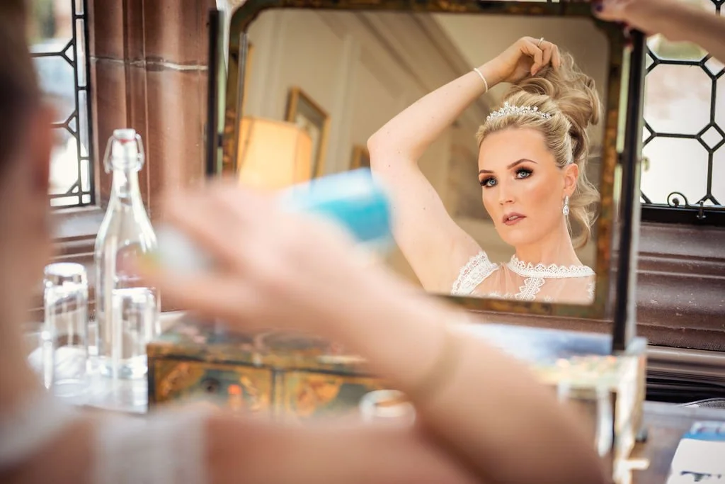 A bride with blonde hair and a tiara looking at herself in a mirror while fixing her hair, with a blurred hand holding a spray bottle in the foreground.