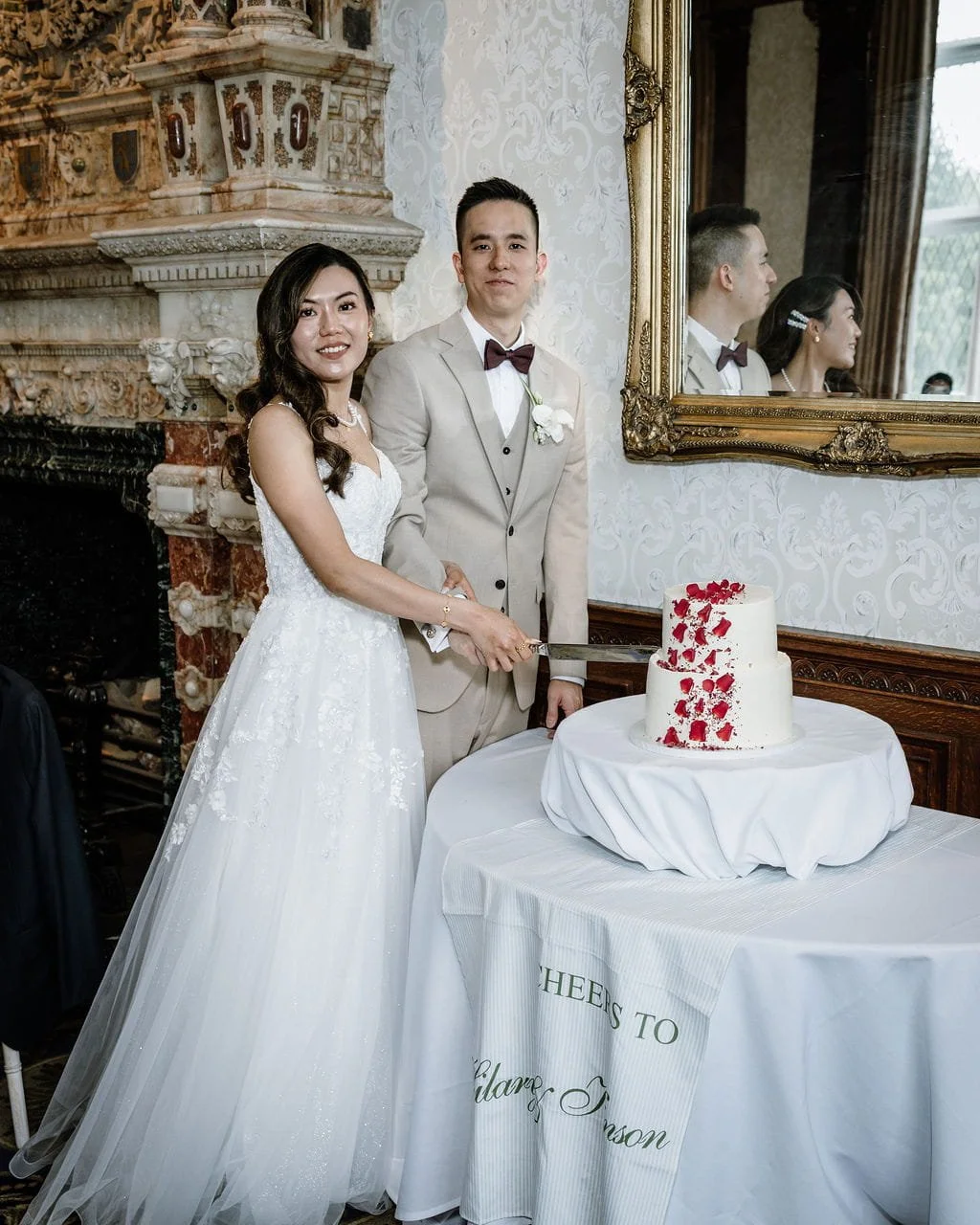 Bride and groom cutting a wedding cake in an elegant room with ornate decor, large mirror, and a table with a white tablecloth.