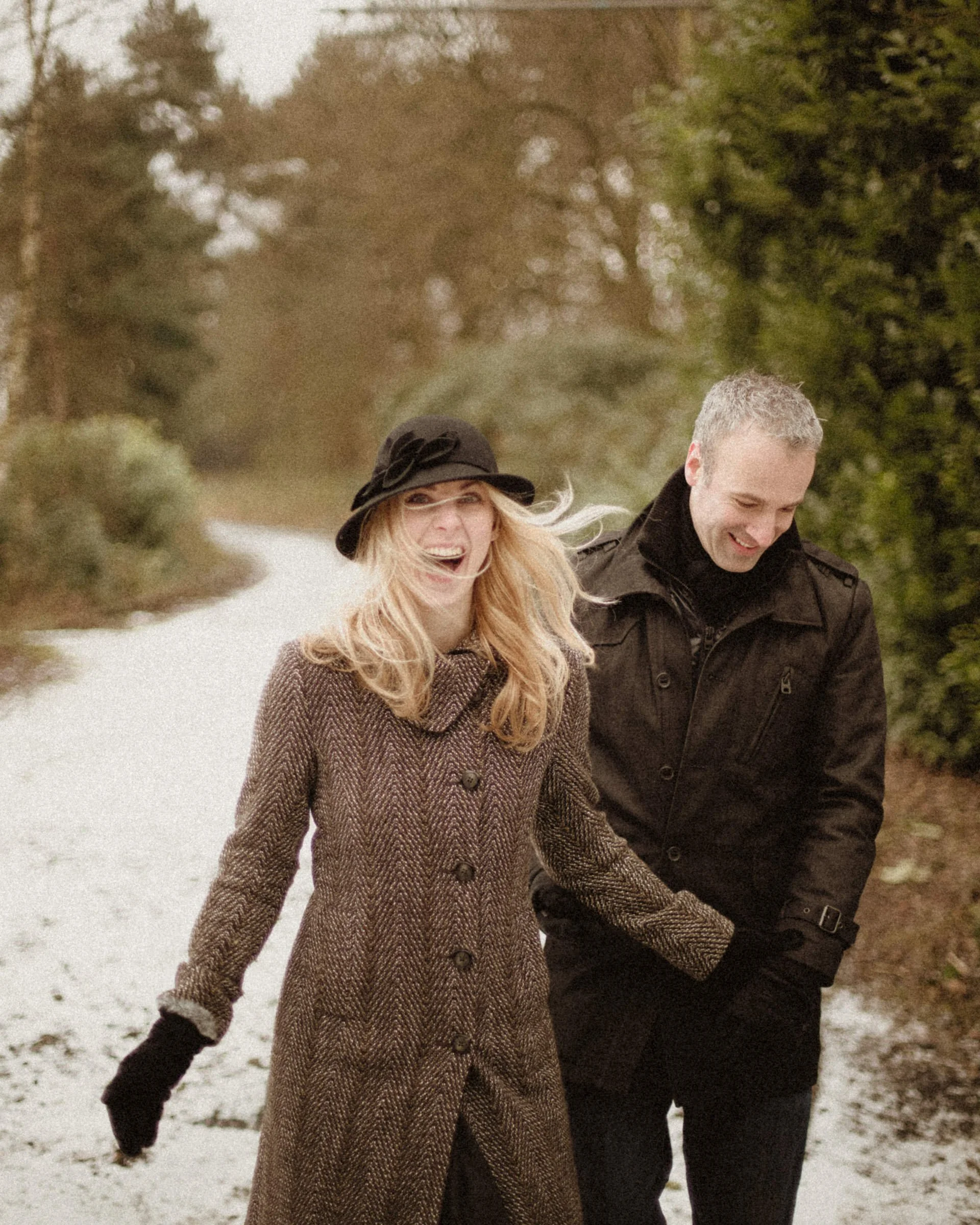 A smiling woman in a brown coat and black hat walking with a man in a black coat along a snowy path surrounded by green trees.