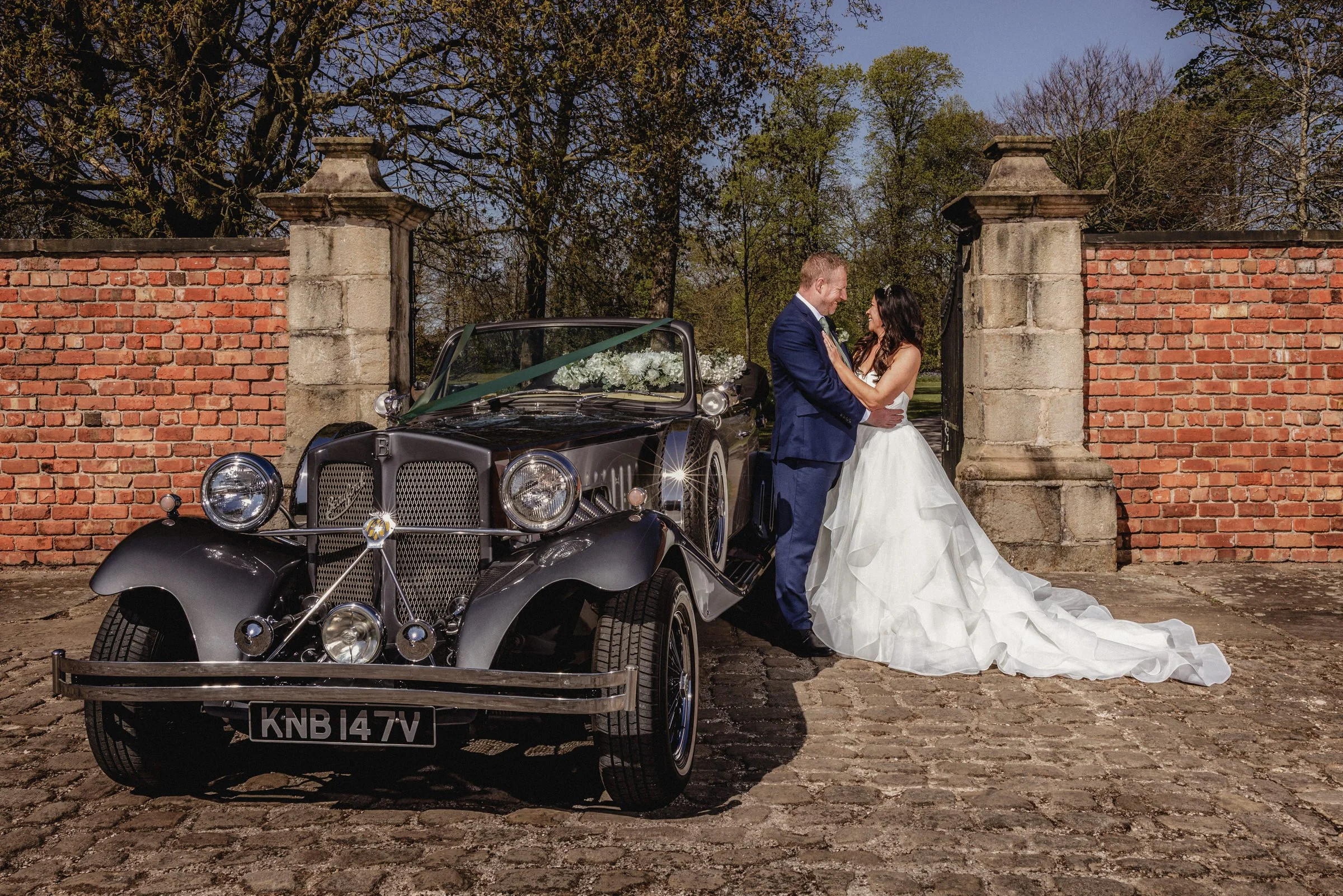 A newlywed couple in wedding attire stands close to each other next to a vintage black car with a floral decoration on the hood, in front of a brick wall with stone pillars, on a cobblestone path, with trees in the background.