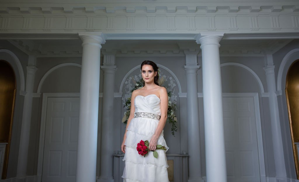 A woman in a white wedding dress holding a bouquet of red roses standing in front of a decorated background with columns and floral arrangements.
