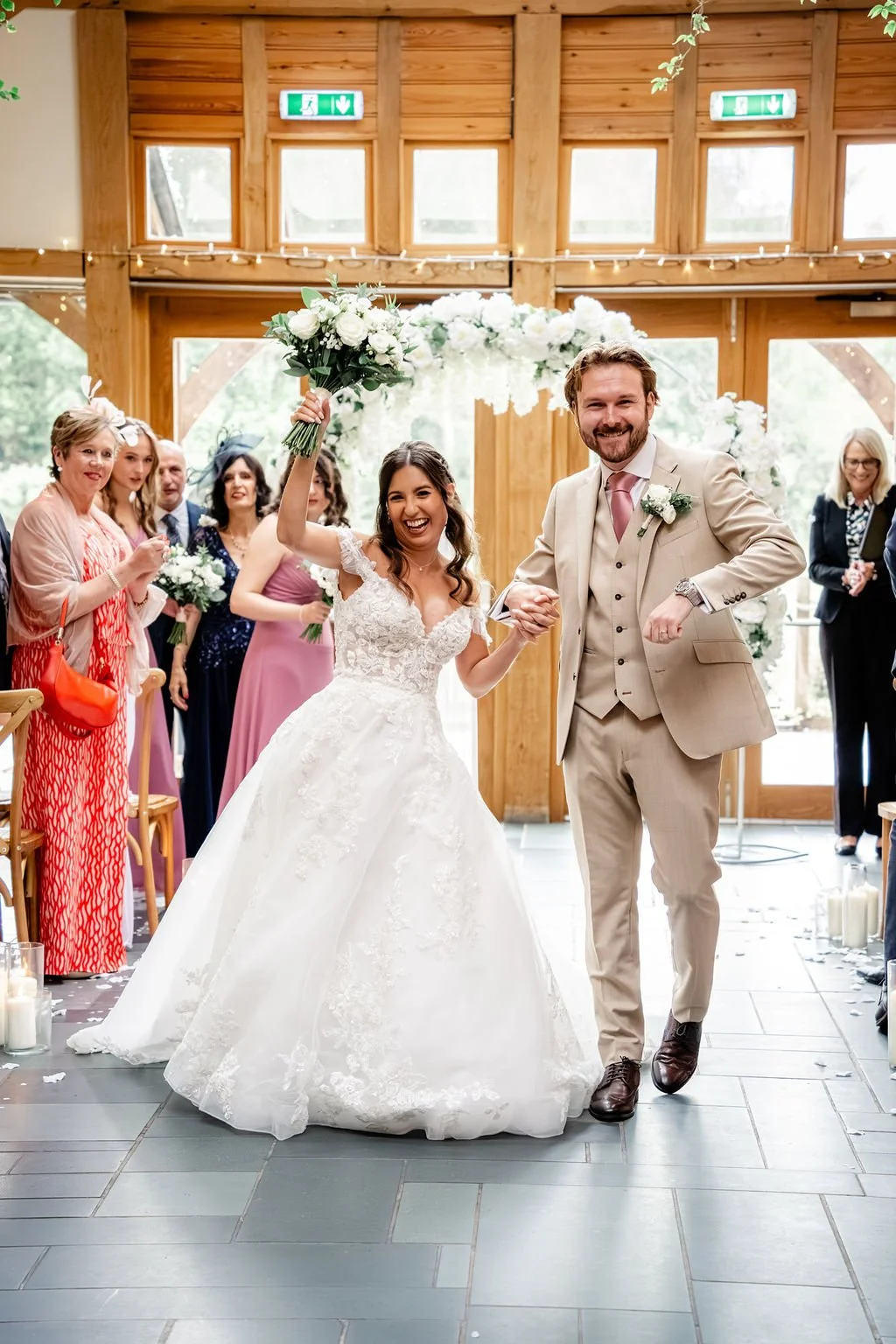 Bride and groom smiling and holding hands, with the bride raising her bouquet, in a wooden hall decorated for a wedding, with guests clapping and taking photos.