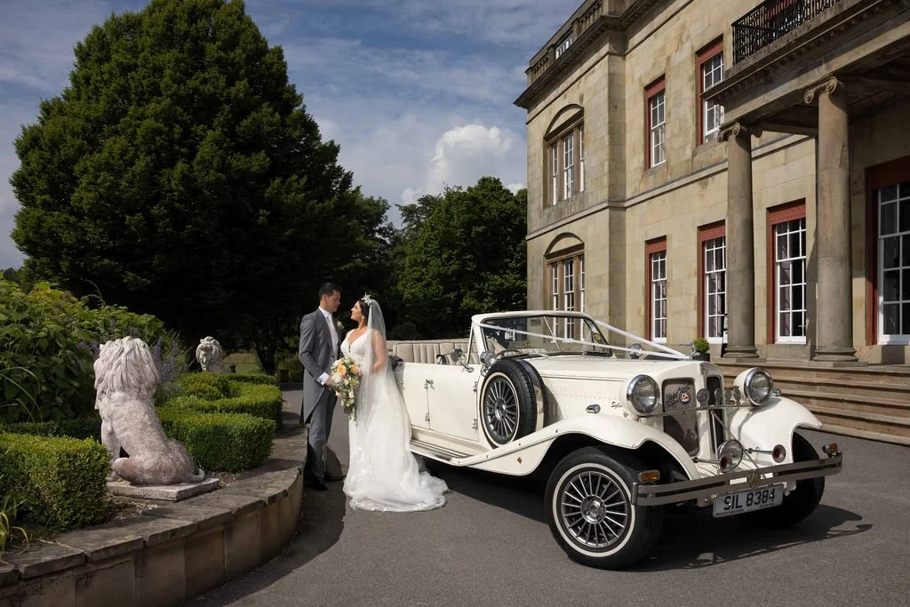 A bride and groom in wedding attire standing next to a vintage white convertible car parked near a stately mansion with columns, steps, and multiple windows, with lush greenery and trees in the background.