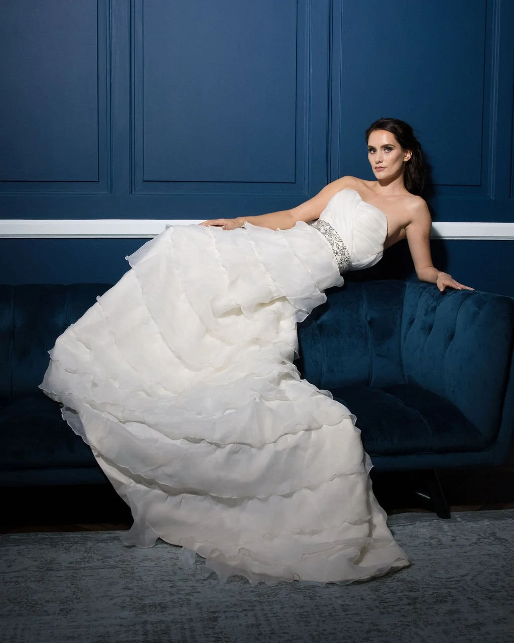 A woman in a strapless white wedding gown lying on a blue velvet couch with dark blue paneled walls behind her.