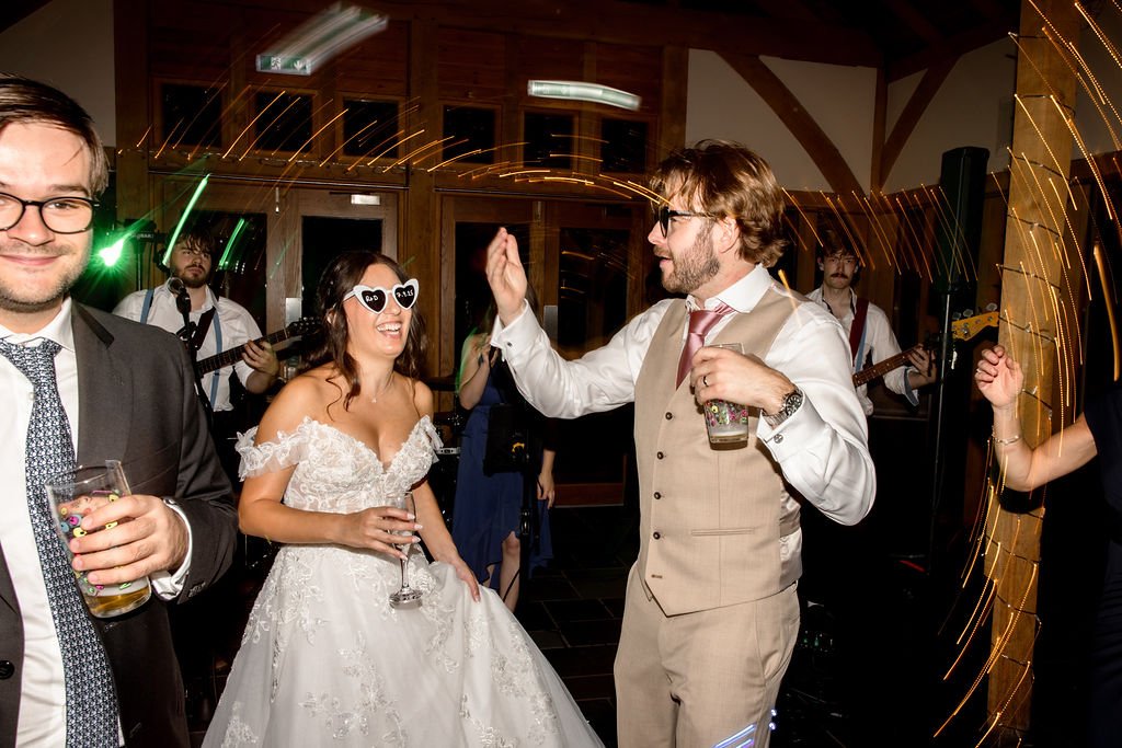 People dancing and celebrating at a wedding reception. A bride in a wedding dress and sunglasses is smiling, while a groom high-fives another man. Guests are holding drinks, and band members are playing guitars in the background. The room has wooden 