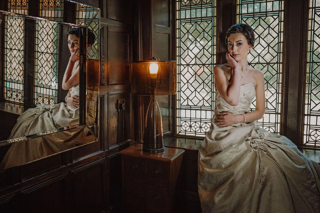 A woman in an elegant strapless gown sitting on a window seat near stained glass windows, with her reflection visible in a nearby mirror, in a dimly lit room with a vintage lamp.