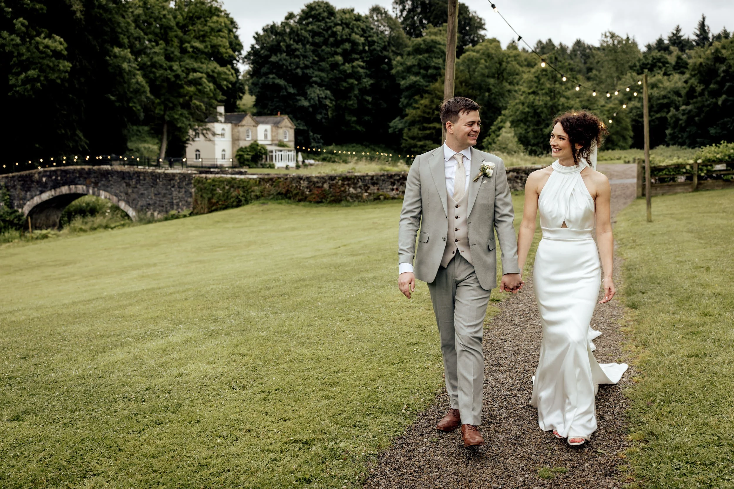 A newlywed couple walking hand in hand on a grassy pathway outdoors, smiling at each other, with a historic stone bridge, white house, and green trees in the background, during the daytime.