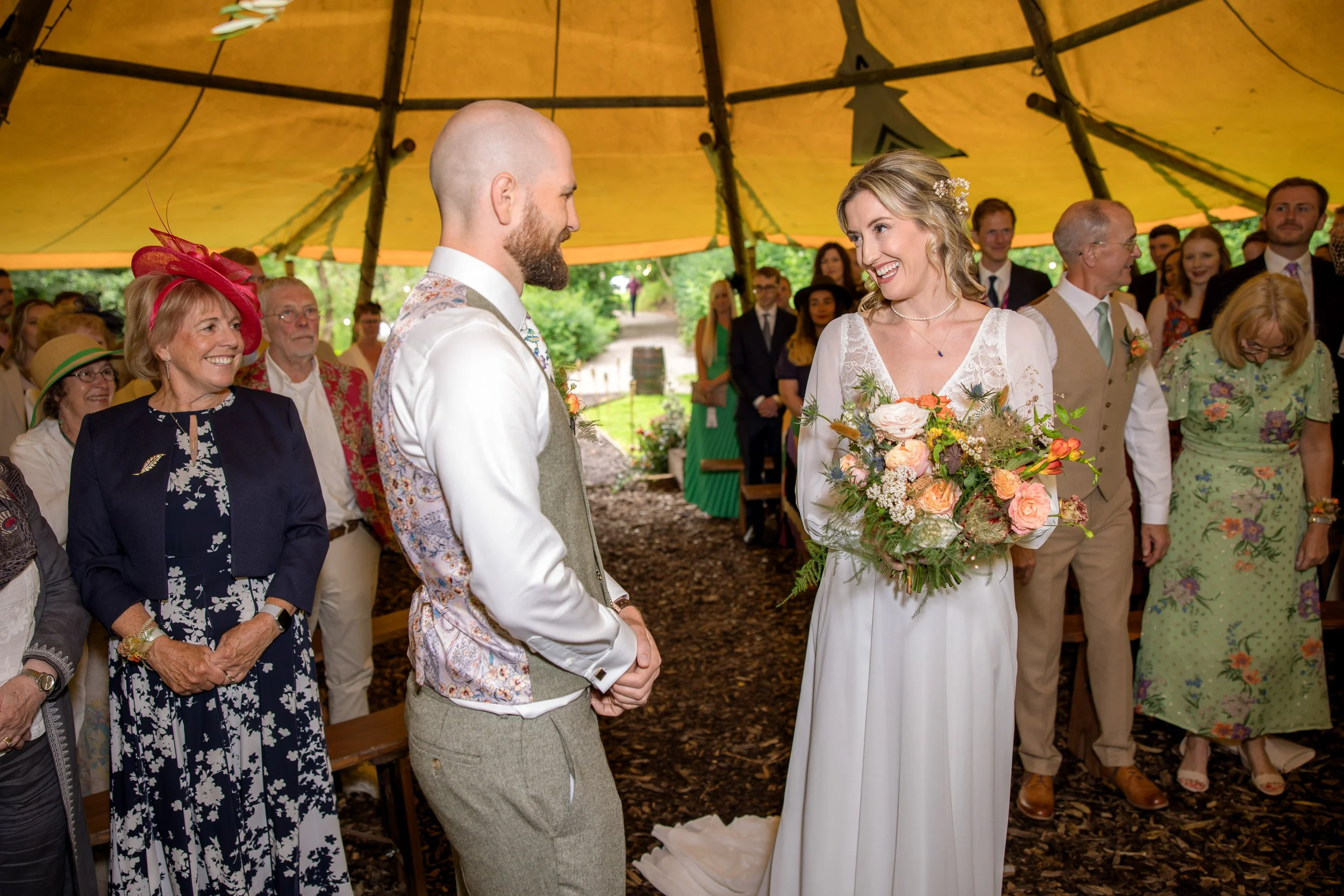A bride and groom standing in a decorated outdoor wedding tent, smiling at each other, with guests watching and smiling in the background.