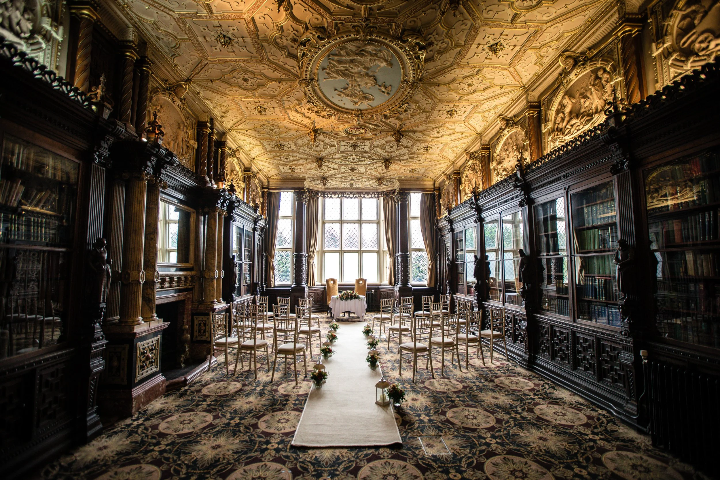An ornately decorated room with high ceilings, large windows, and intricate ceiling moldings. The room is set up for a wedding ceremony with a white aisle runner, floral arrangements, and two chairs at the front. The walls are lined with dark wooden bookshelves and the floor has a patterned carpet.