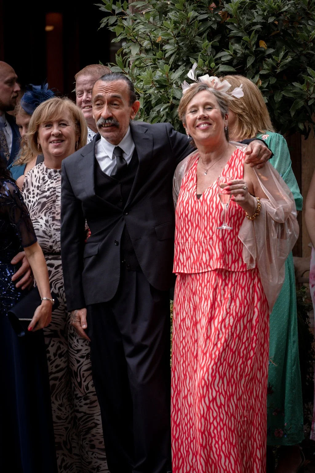 Group of well-dressed people at a social gathering, smiling, with two women in the front holding a glass of champagne, one wearing a pink patterned dress with a floral headpiece and the other in formal attire.