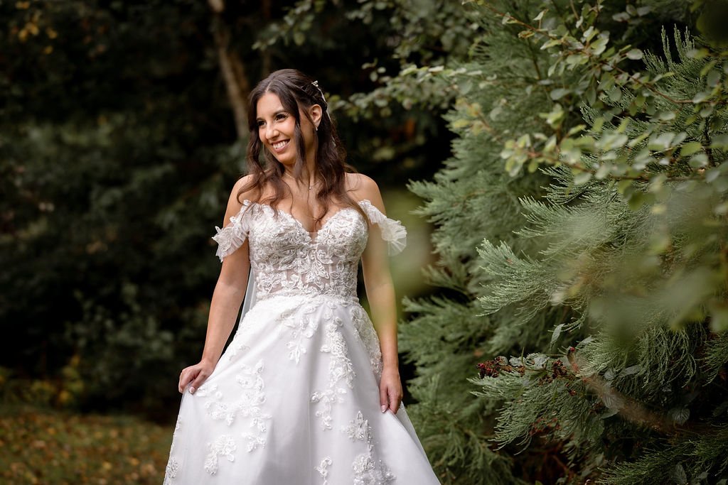 A woman in a white wedding dress standing outdoors near trees and foliage, smiling.