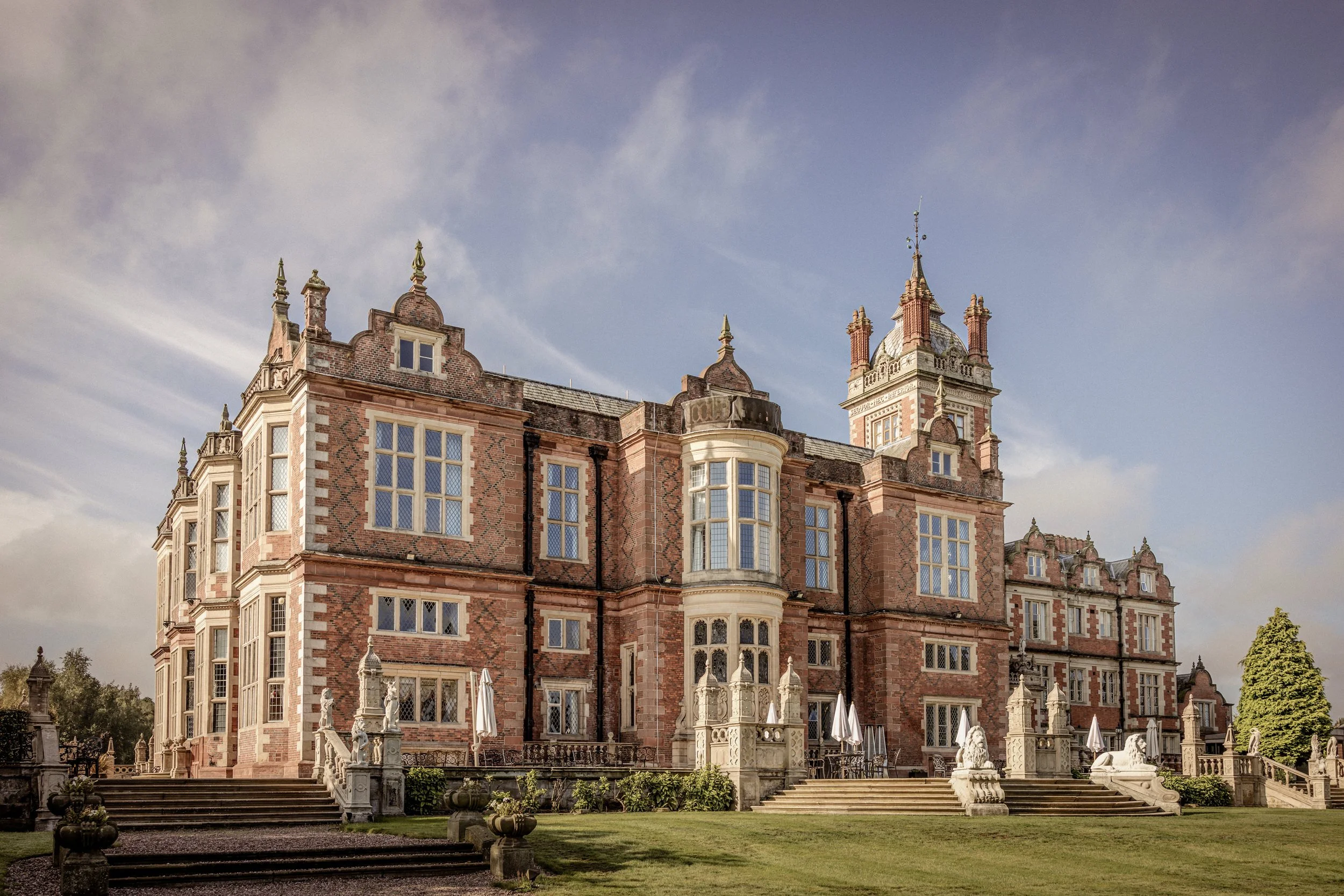 A grand historic castle with red brick and stone details, multiple towers, large windows, and decorative architectural elements, set against a partly cloudy sky with greenery around.