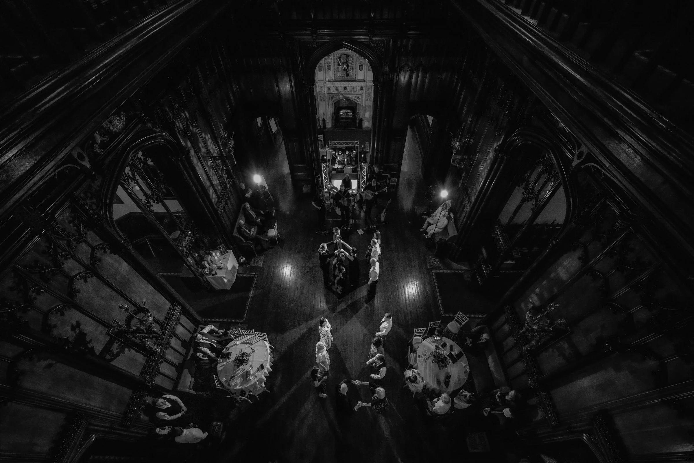 Black and white aerial view of an indoor wedding reception with guests dancing, seated at tables, and musicians performing on a stage at the far end of the ornate, wood-paneled room.