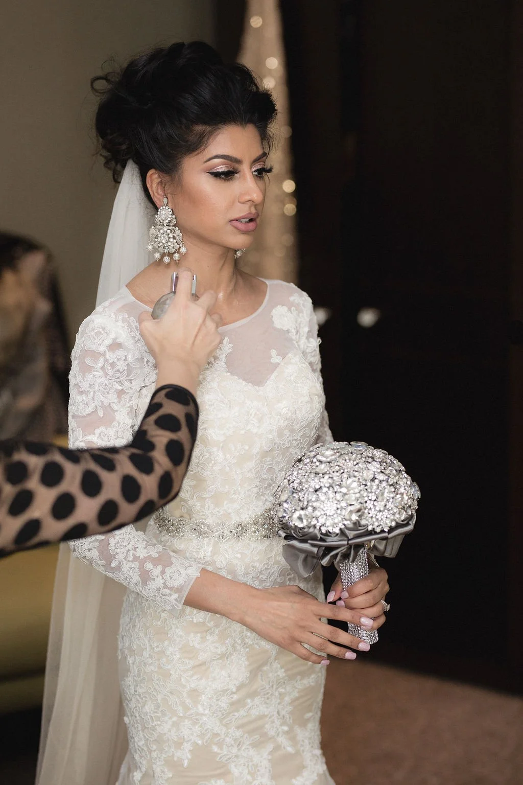 A bride with dark hair styled up, wearing wedding earrings and a lace wedding dress, holding a bouquet of rhinestones and fabric, getting her makeup done.