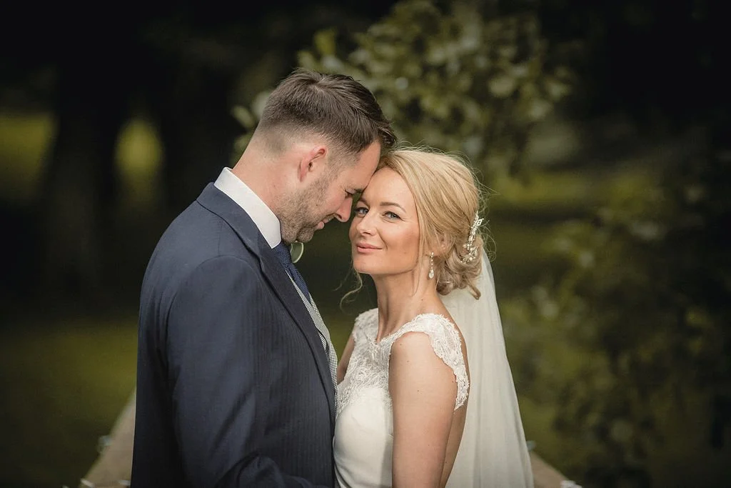 A bride and groom sharing a moment outdoors, with the groom leaning his forehead against the bride's, against a background of blurred trees and greenery.