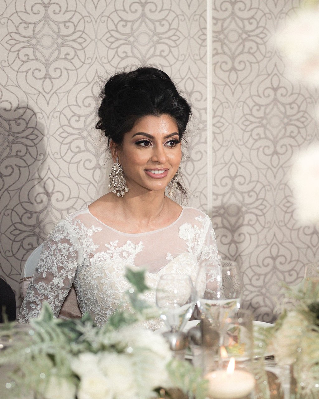 A smiling woman with dark hair styled in an updo, wearing white lace bridal dress and large chandelier earrings, seated at a table with floral arrangements, candles, and glassware, with a patterned wallpaper background.