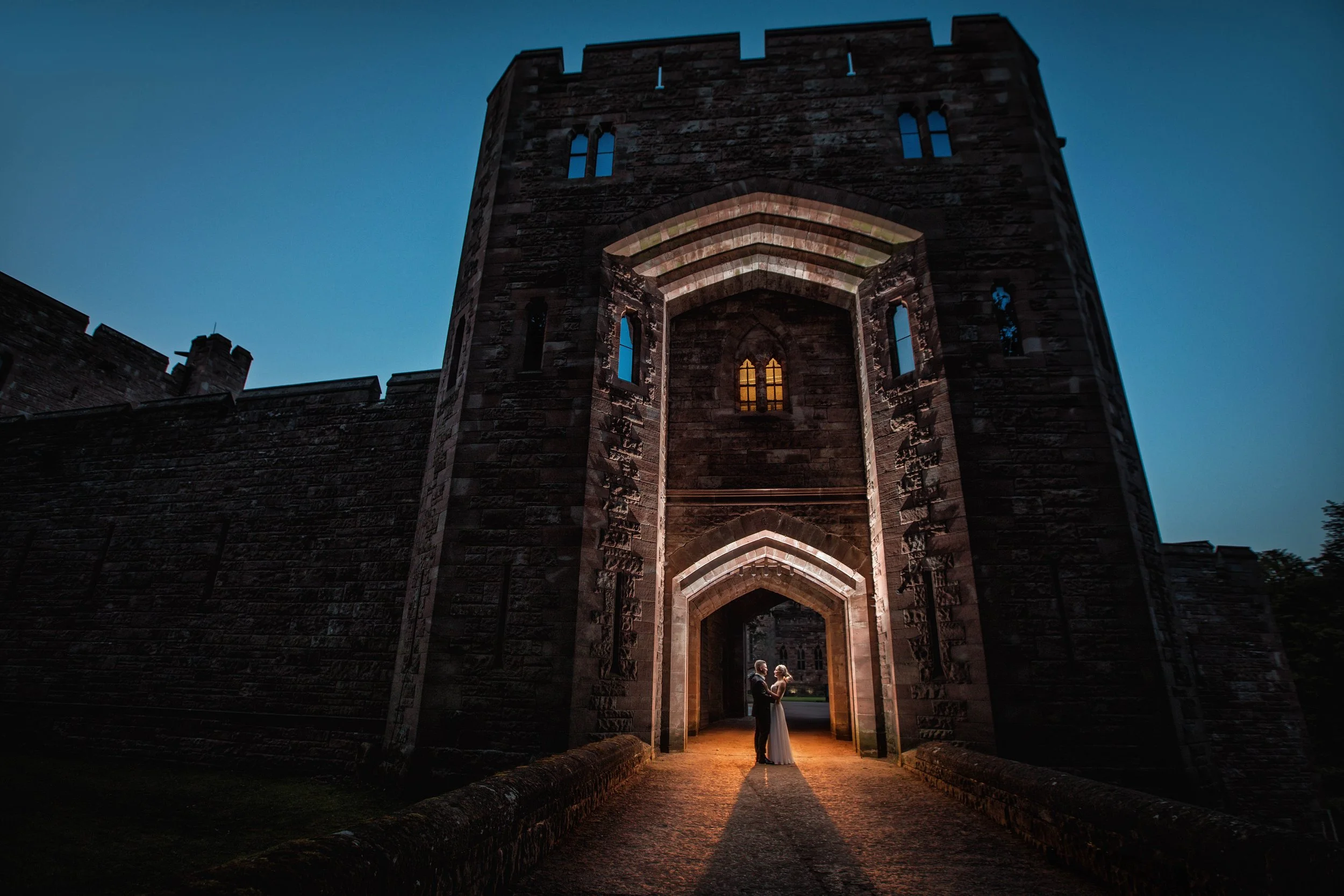 Couple standing and embracing under an arched medieval castle gate during dusk with lit windows and fading daylight in the background.