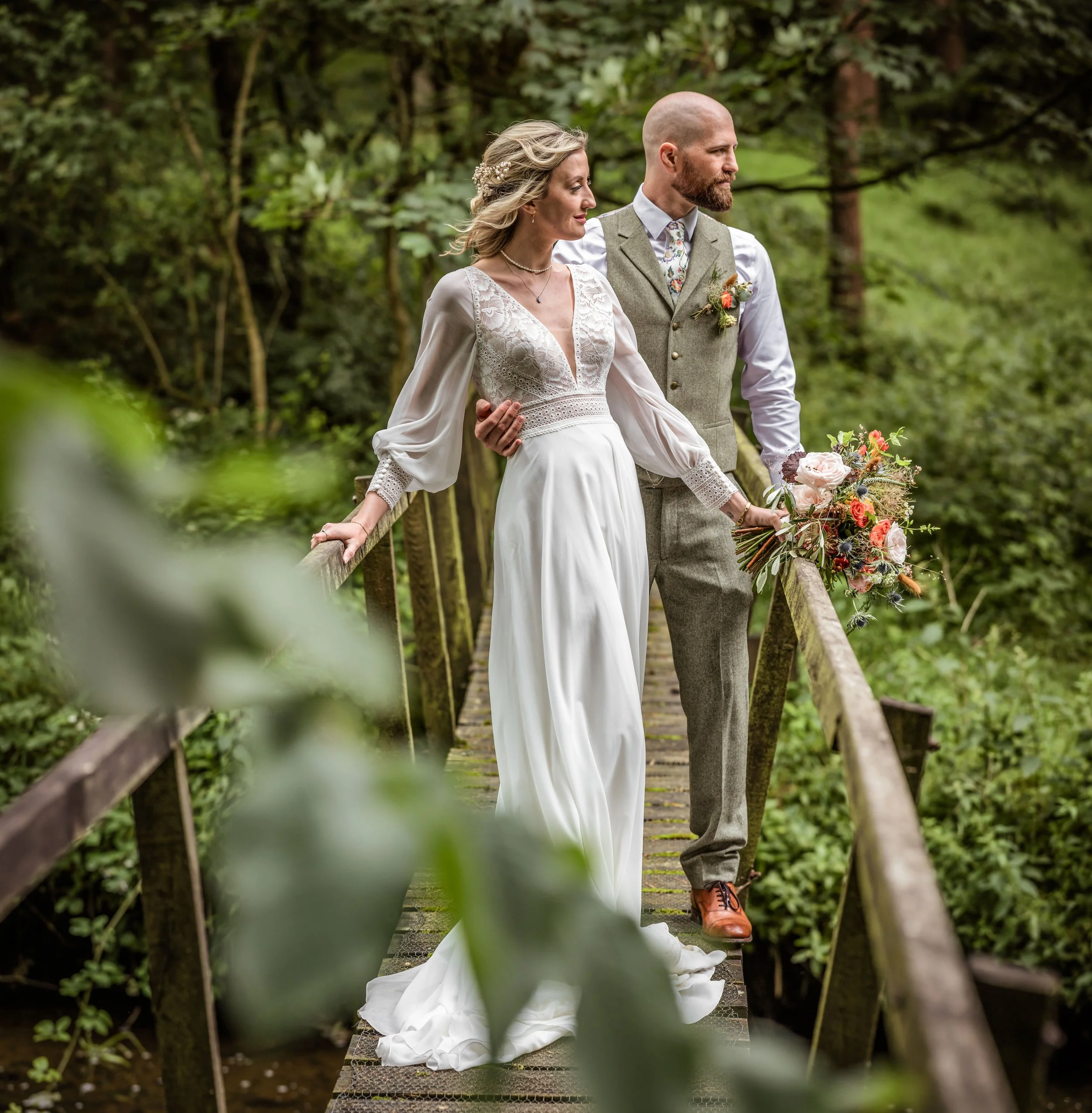 A bride and groom holding hands on a wooden bridge in a wooded area, with the bride holding a bouquet of flowers.
