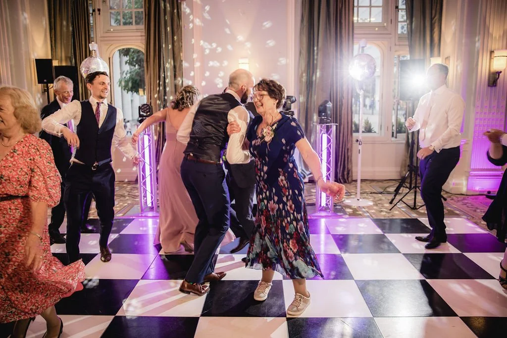 People dancing and enjoying themselves at a wedding reception or party in a decorated ballroom with checkered black and white floor, purple lighting, and large windows.