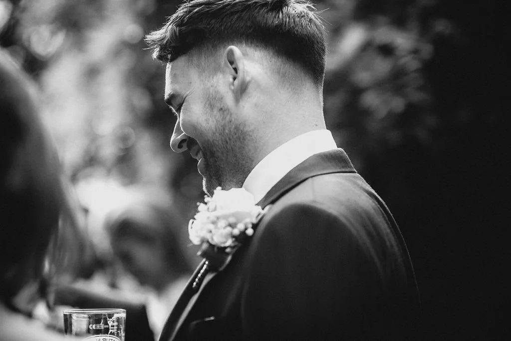 A man in formal attire, smiling with eyes closed, at a wedding or celebration event, with blurred background.