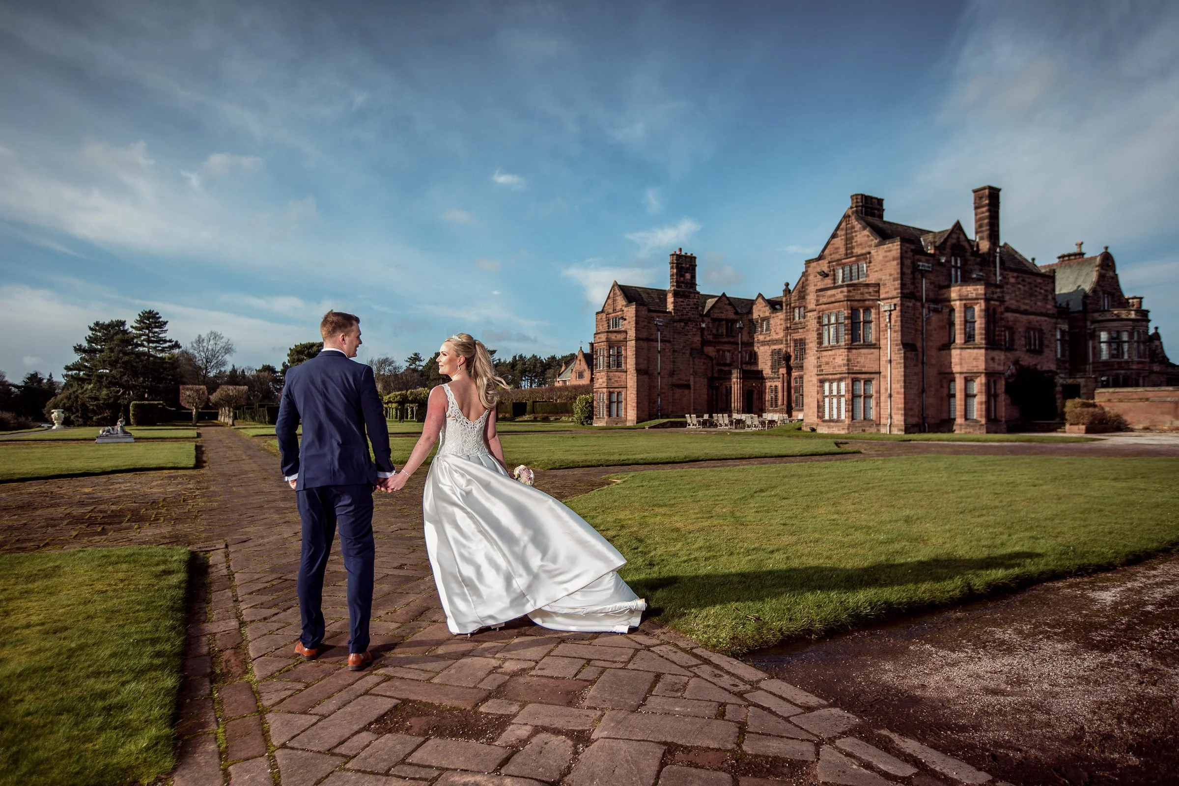 Bride and groom walking hand in hand on brick pathway outside large historic mansion under blue sky