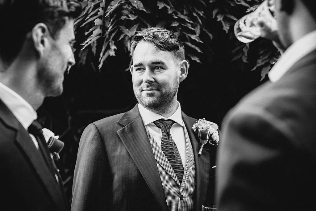 A groom in a suit and tie standing with two men in suits during a wedding ceremony or reception, in an outdoor setting with lush greenery background.