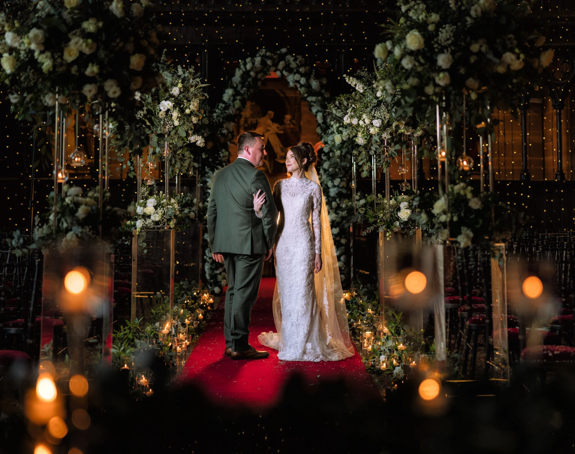 A bride and groom stand close together under a floral arch on a decorated aisle during their wedding at night, surrounded by candles and fairy lights.
