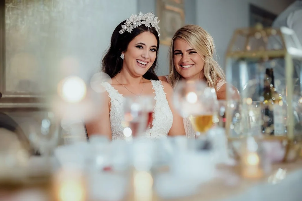 Two women smiling at a table, one wearing a wedding dress and a floral headpiece, the other in a light dress, in a decorated indoor setting with glasses of beer and gold accents.