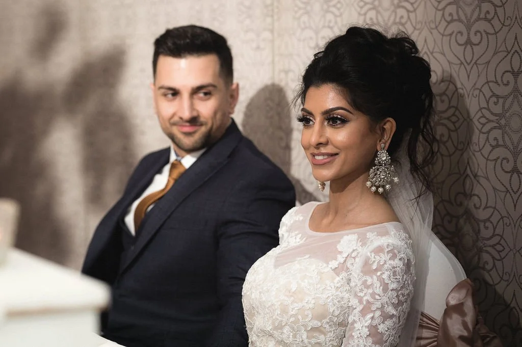 A bride and groom sitting side by side, smiling, at their wedding. The bride wears a lace wedding dress and large earrings, while the groom is dressed in a dark suit with a white shirt and a brown tie.