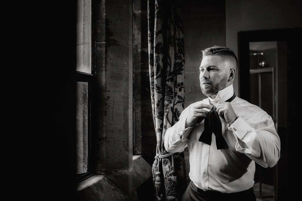 A man in a white dress shirt adjusts a black bow tie in front of a window with patterned curtains, in a black and white photo.
