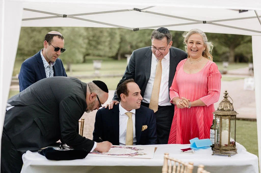 A group of people gathered around a table outdoors during daytime, with a young man signing a document, others smiling and looking on, under a white canopy with trees in the background.