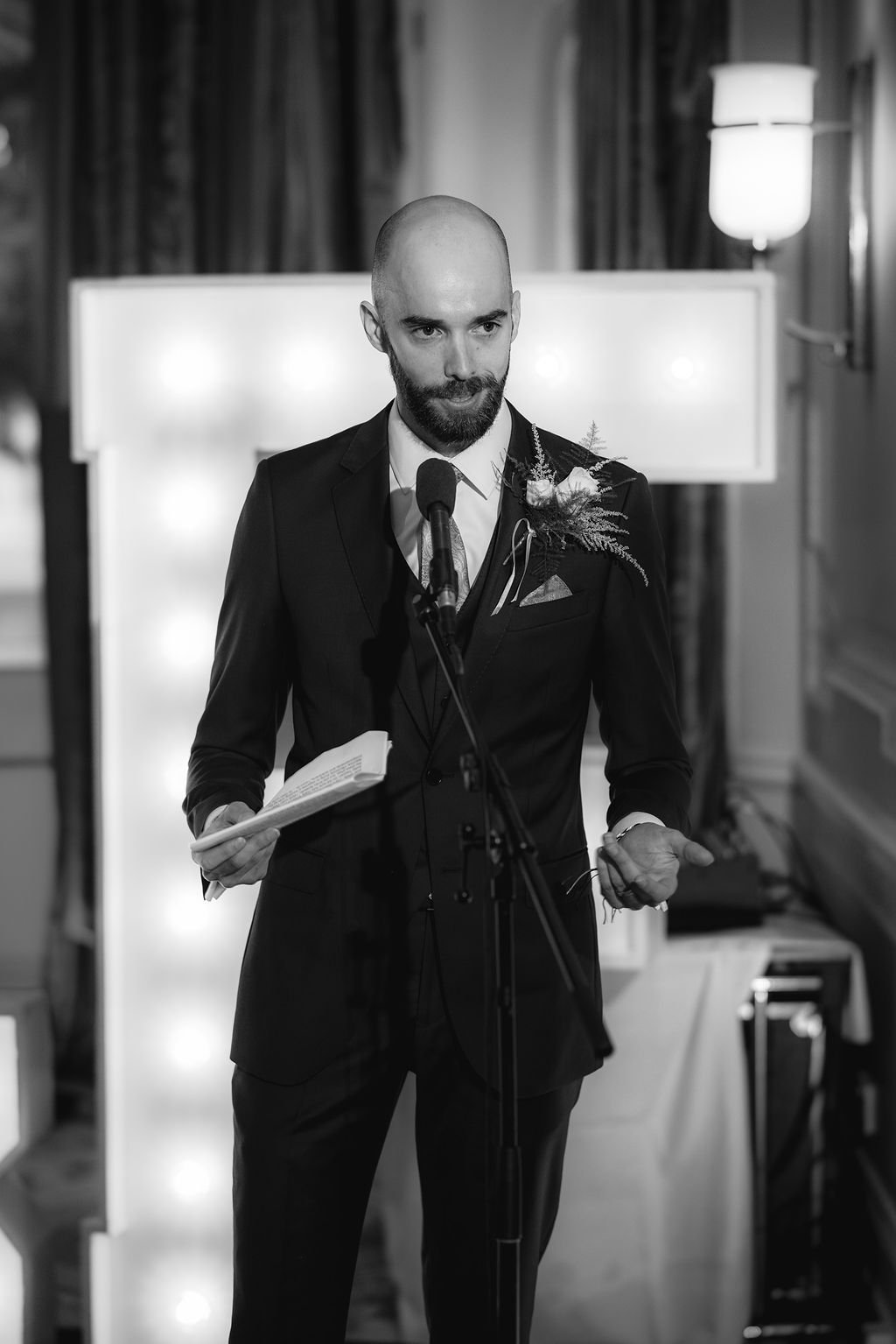 A man with a shaved head and beard in a suit giving a speech at a formal event, standing in front of a microphone, holding papers, with a flower boutonniere on his lapel.