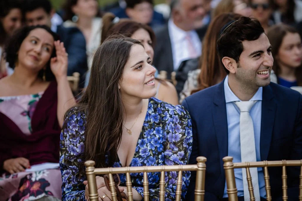 An audience seated on gold chairs at an outdoor event, with a focus on a young woman in a blue floral dress and a young man in a blue suit and white tie, both smiling.