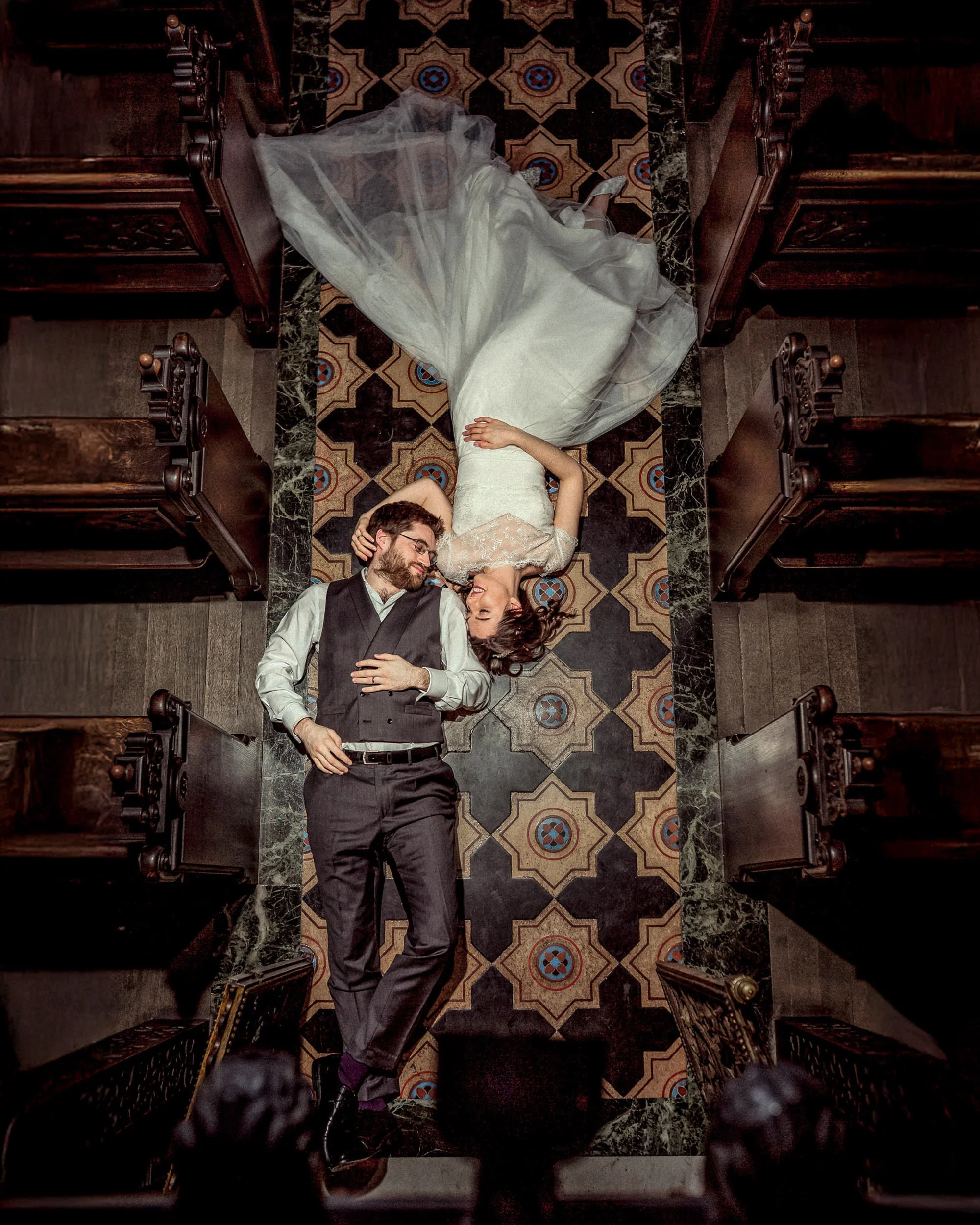 A bride and groom lying face to face on a patterned church aisle floor, holding hands and gazing at each other, surrounded by empty wooden pews.