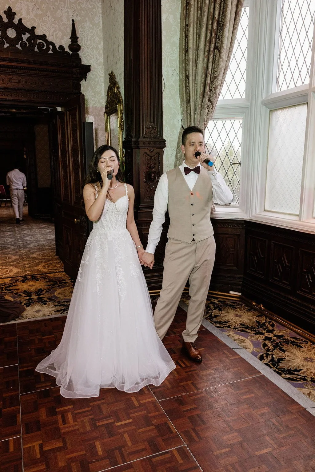 Bride and groom holding hands and singing into microphones during their wedding reception in a decorated hall with patterned wallpaper, large windows with curtains, and dark wood paneling.