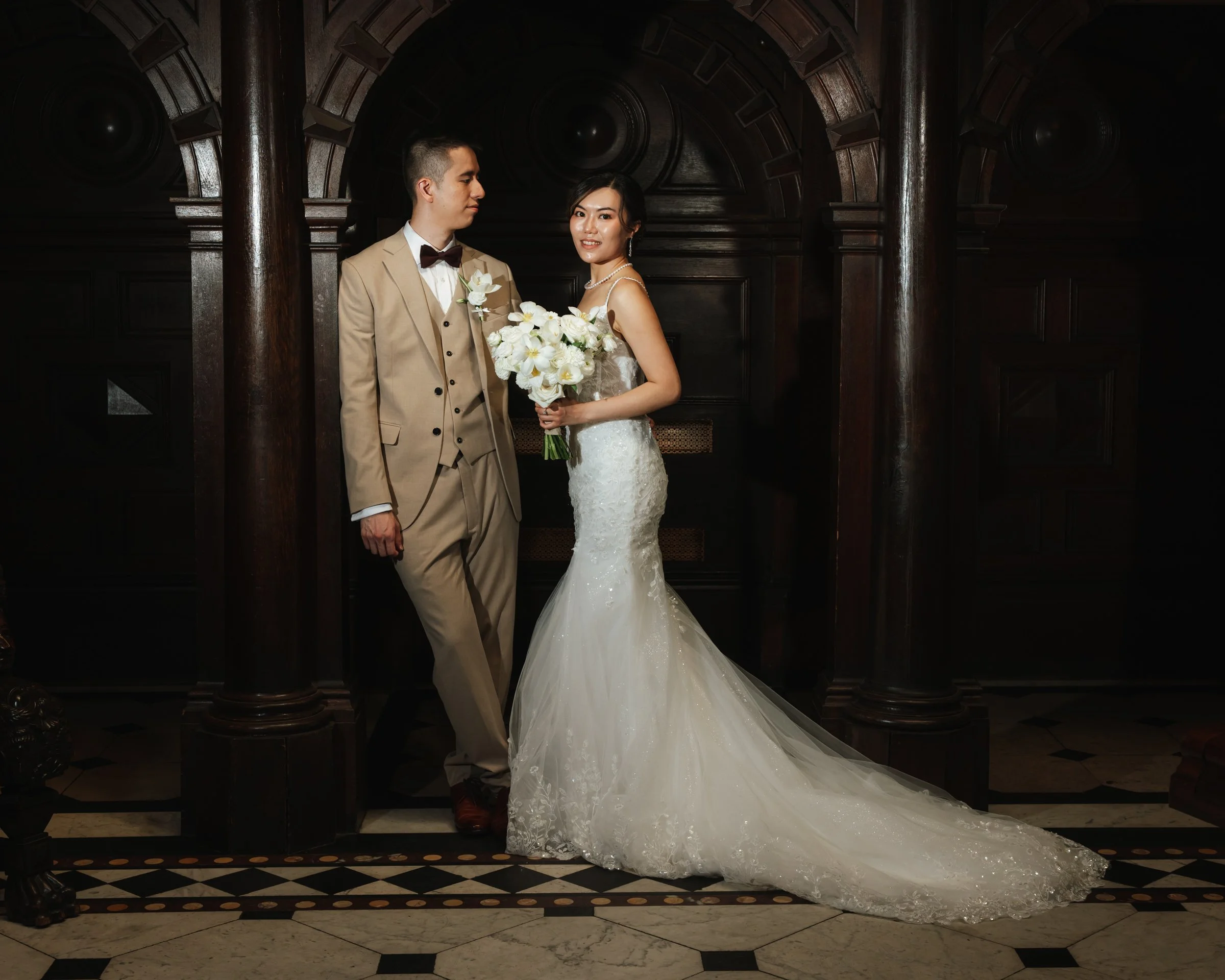 A bride and groom standing in front of a dark wooden backdrop, with the bride holding a bouquet of white flowers. The groom is wearing a tan suit with a bow tie, and the bride is in a white, lace wedding gown with a pearl necklace and earrings. The b