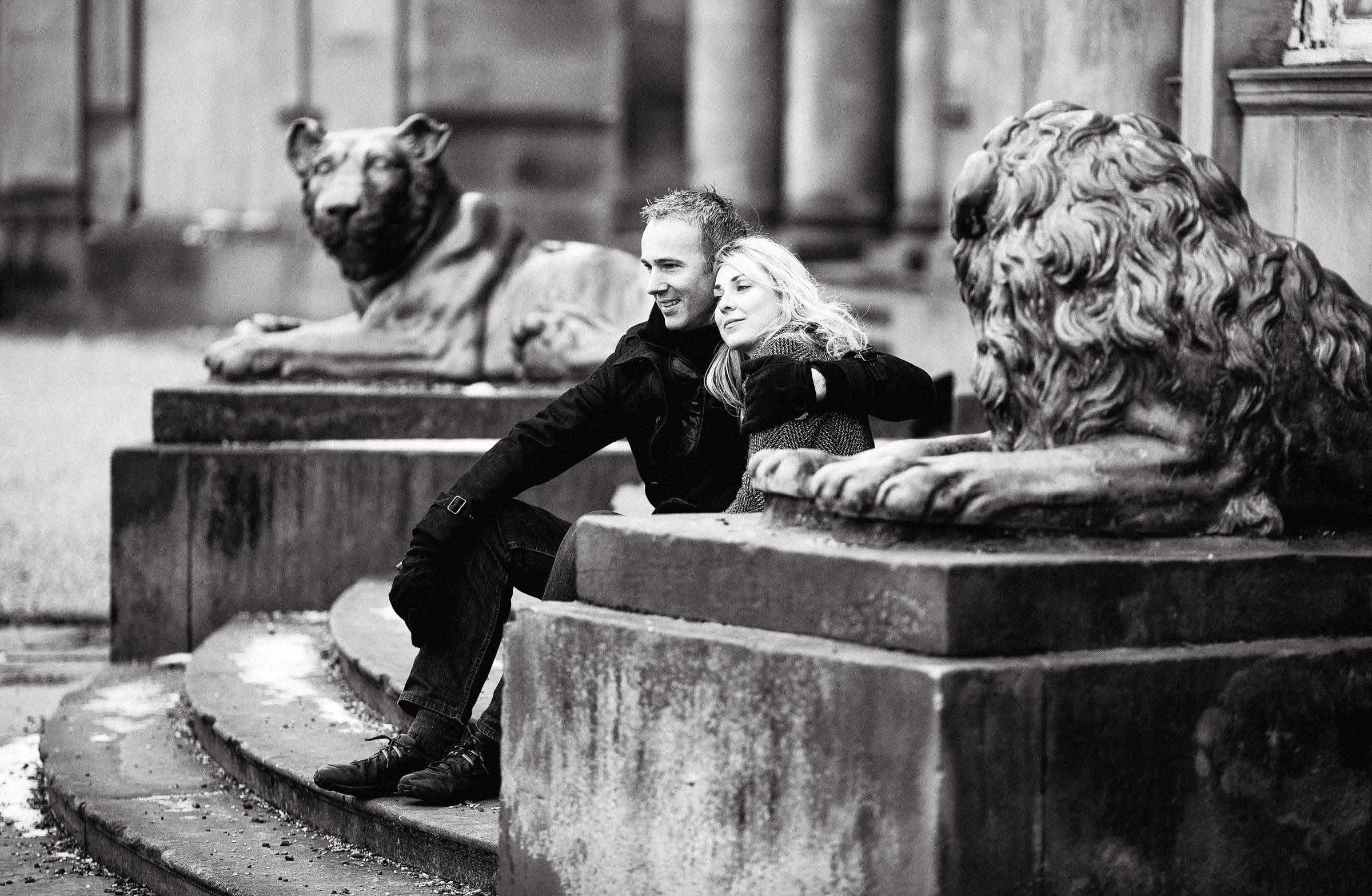 A man and woman sitting on a stone ledge between two lion statues made of stone in an outdoor urban setting, captured in black and white.