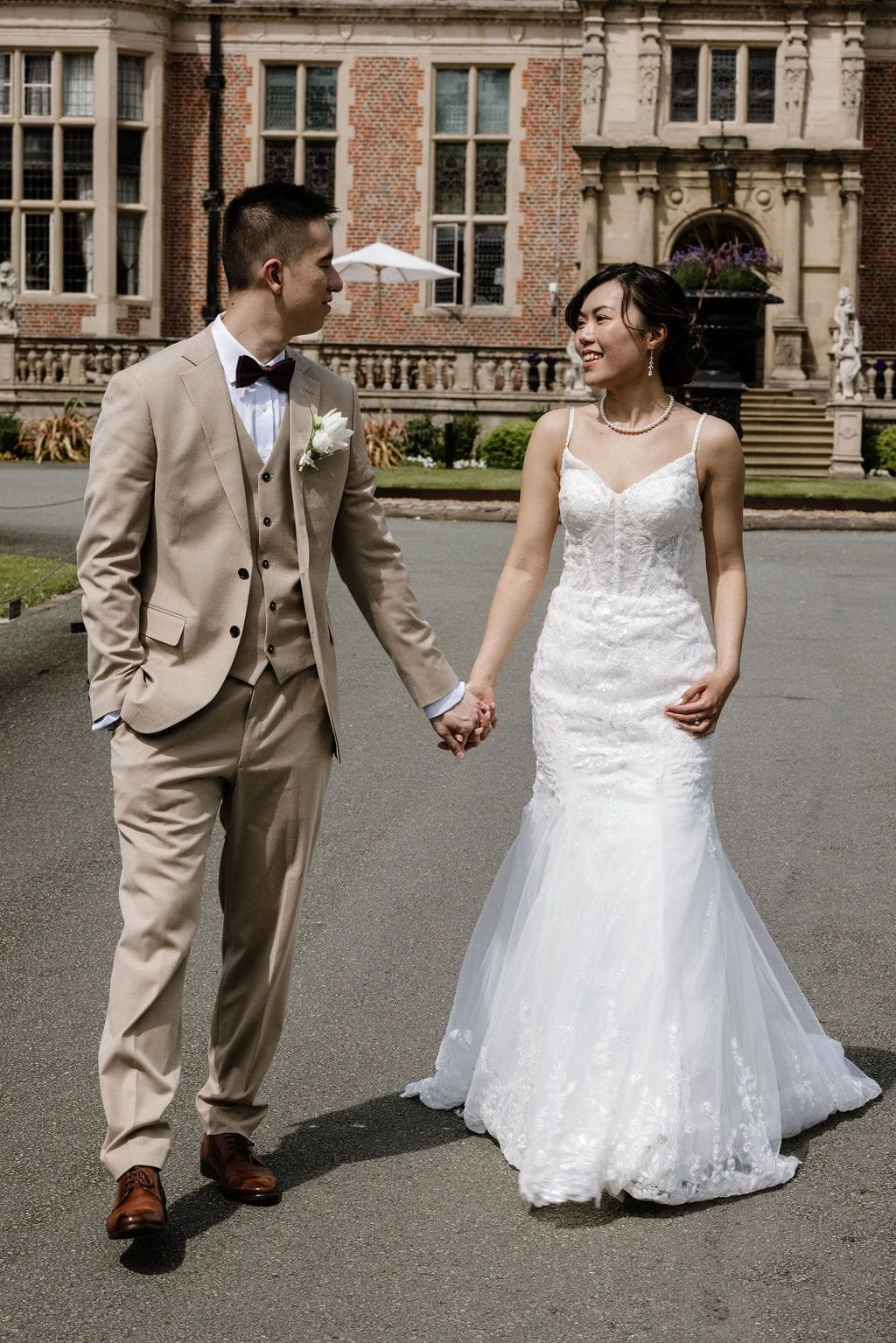 A bride and groom holding hands outside a large brick and stone mansion. The bride wears a white lace wedding dress and pearl jewelry, and the groom wears a tan suit with a white shirt, burgundy bow tie, and boutonniere. They smile at each other.