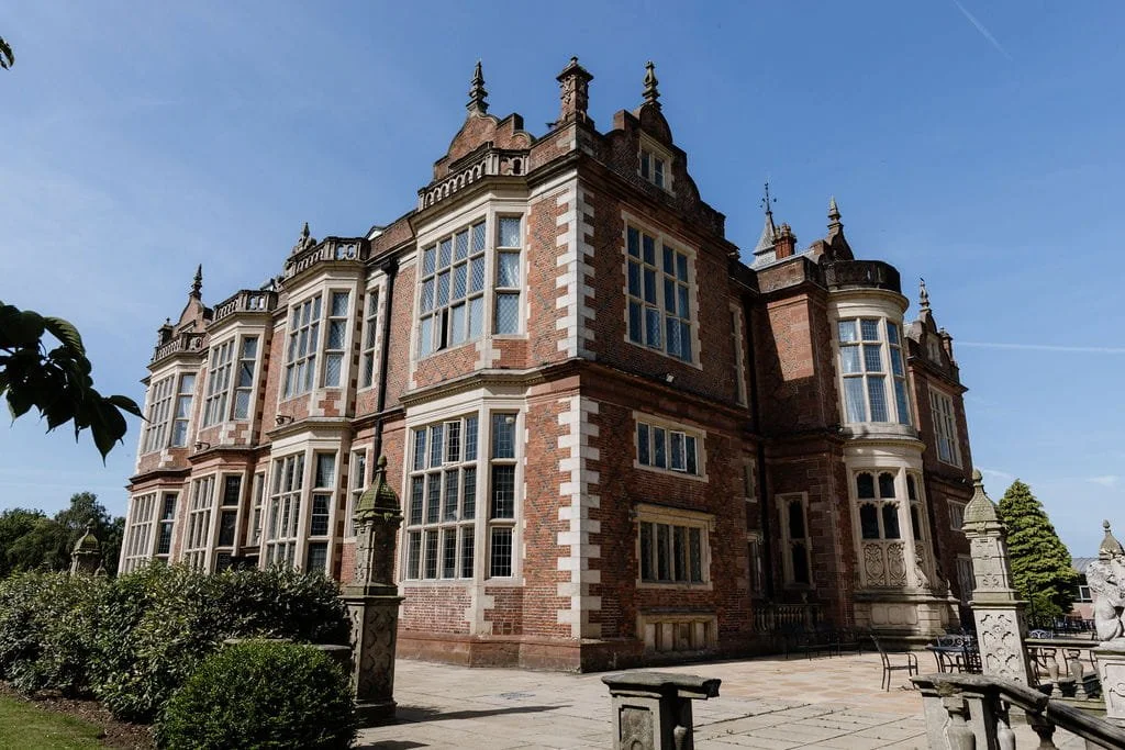 Large historic red brick castle with intricate architectural details, multiple large windows, and stone accents, surrounded by a garden and outdoor furniture under a clear blue sky.