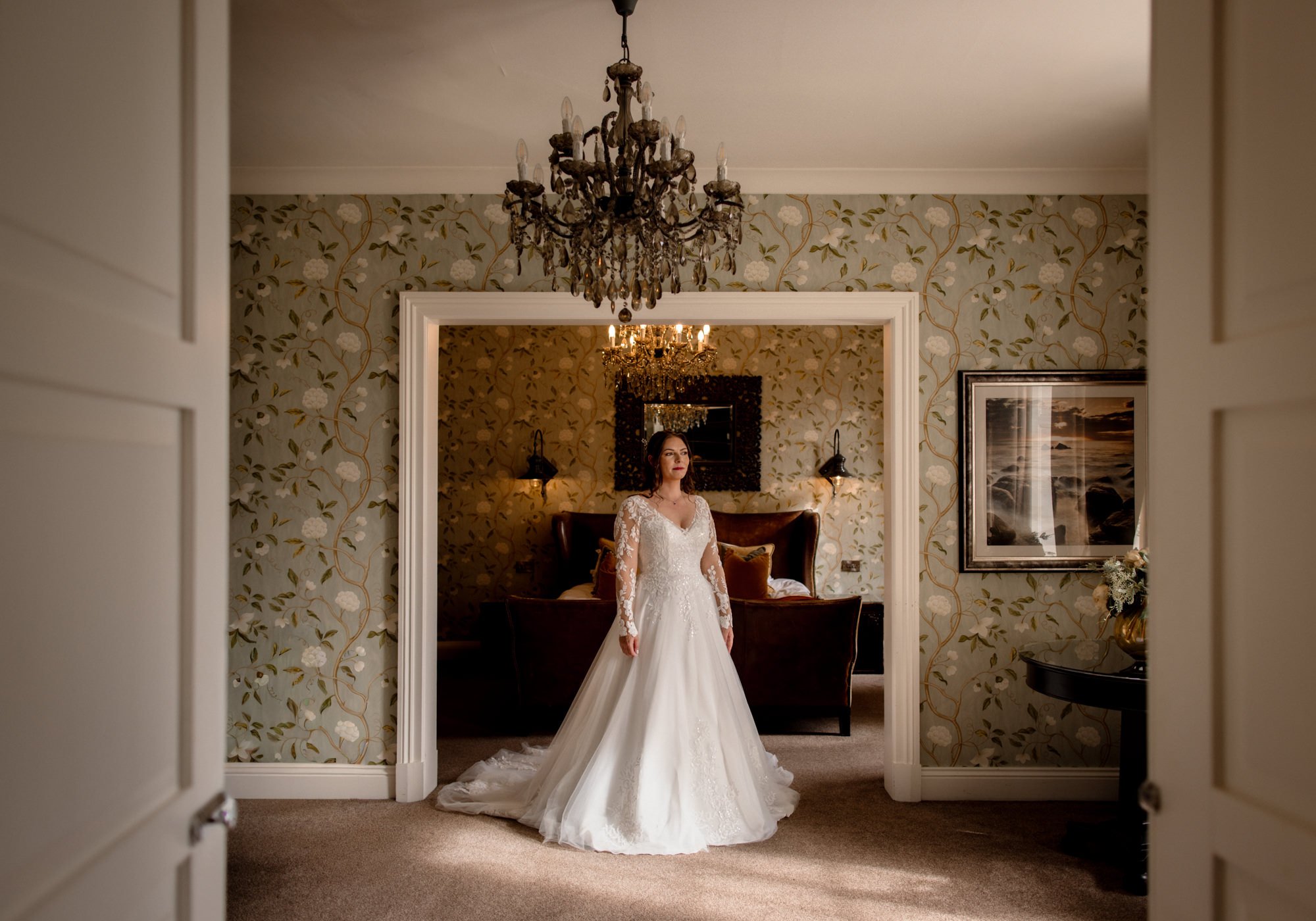 A woman in a white wedding gown standing in a vintage hotel room with floral wallpaper, chandelier, and art decoration.