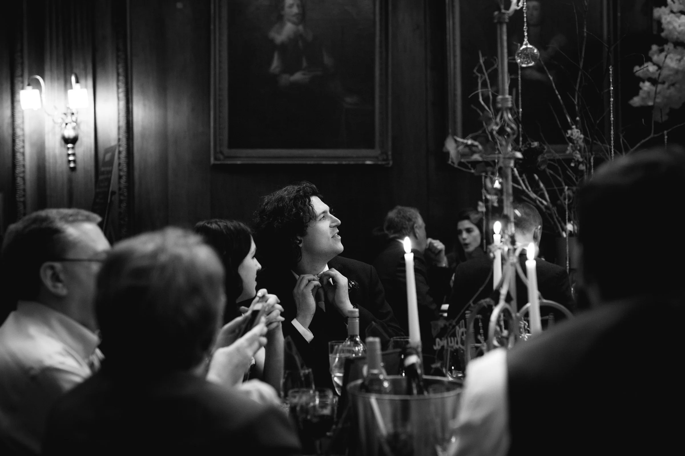 People sitting at a dinner table in a decorated room with candlelight, engaged in conversation and looking towards a focal point.