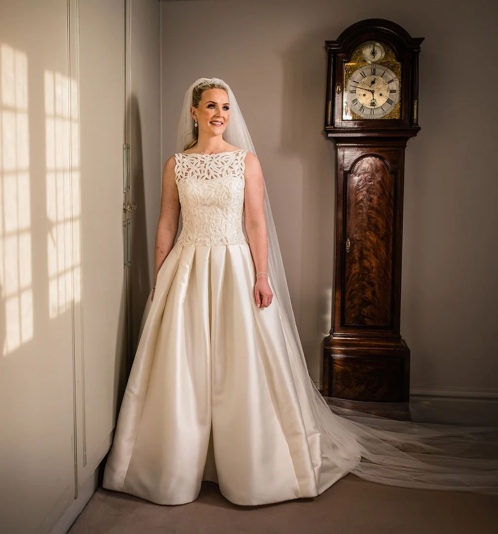 Bride in a wedding dress standing beside a wooden grandfather clock and a wall with window shadows.