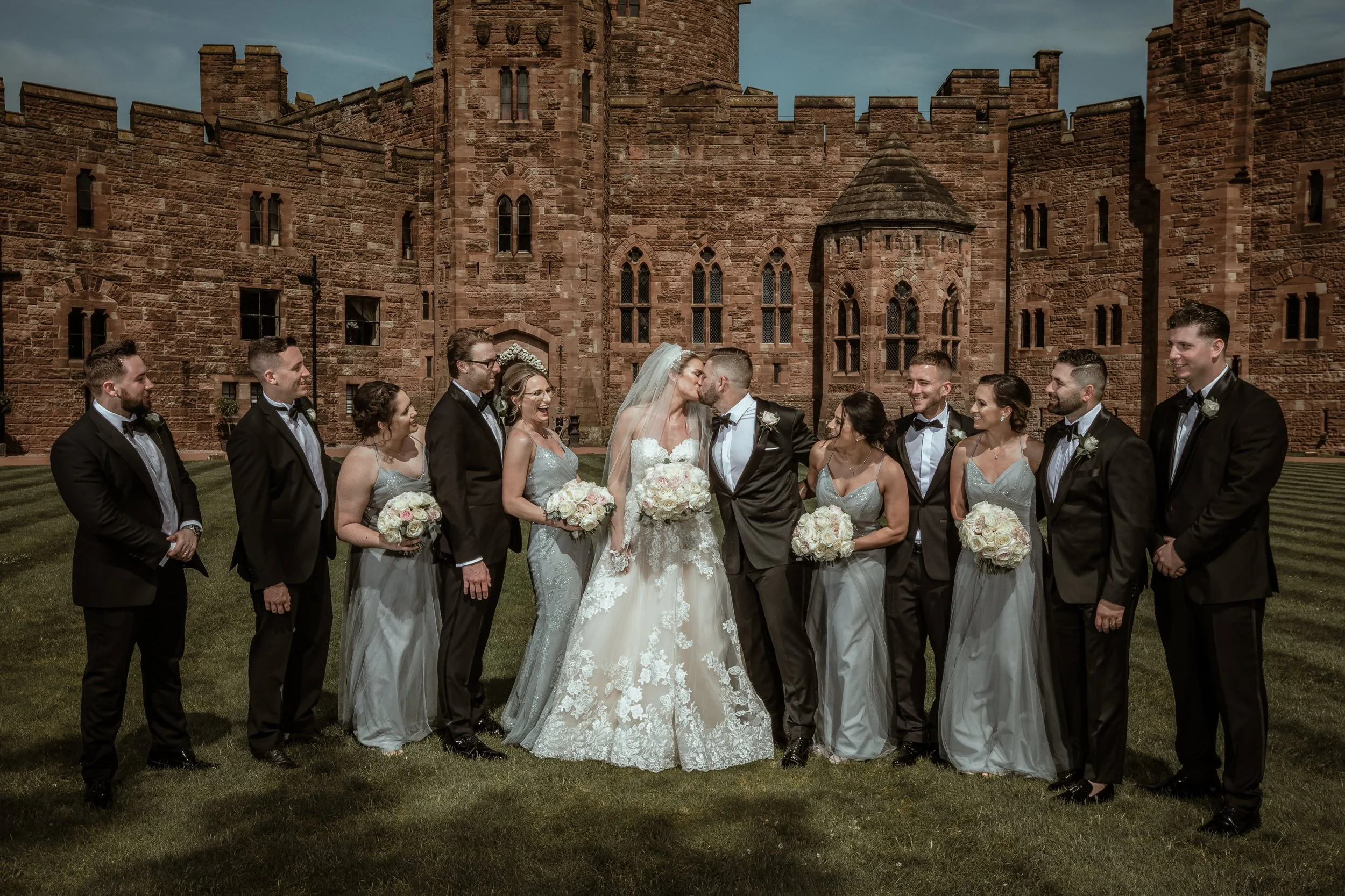 A wedding party standing outside a historic castle, with the bride and groom kissing in the center surrounded by bridesmaids and groomsmen in formal attire, all holding bouquets of flowers.