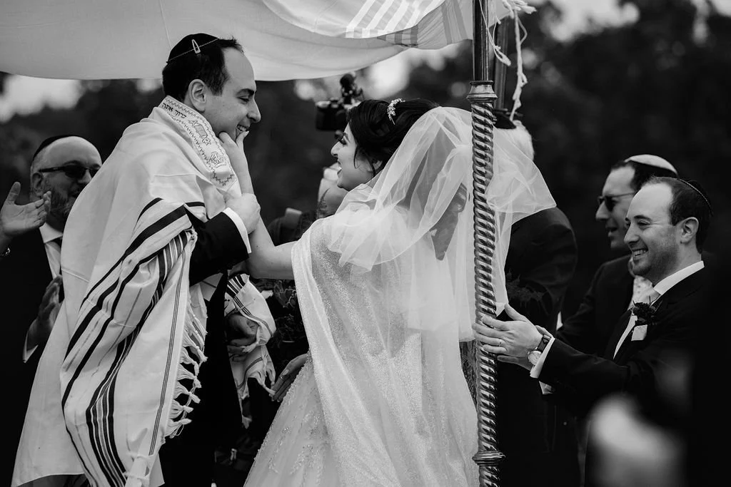 A bride and groom smile at each other during a Jewish wedding ceremony, with the bride holding the groom's face and the groom touching his chin, surrounded by friends and family.