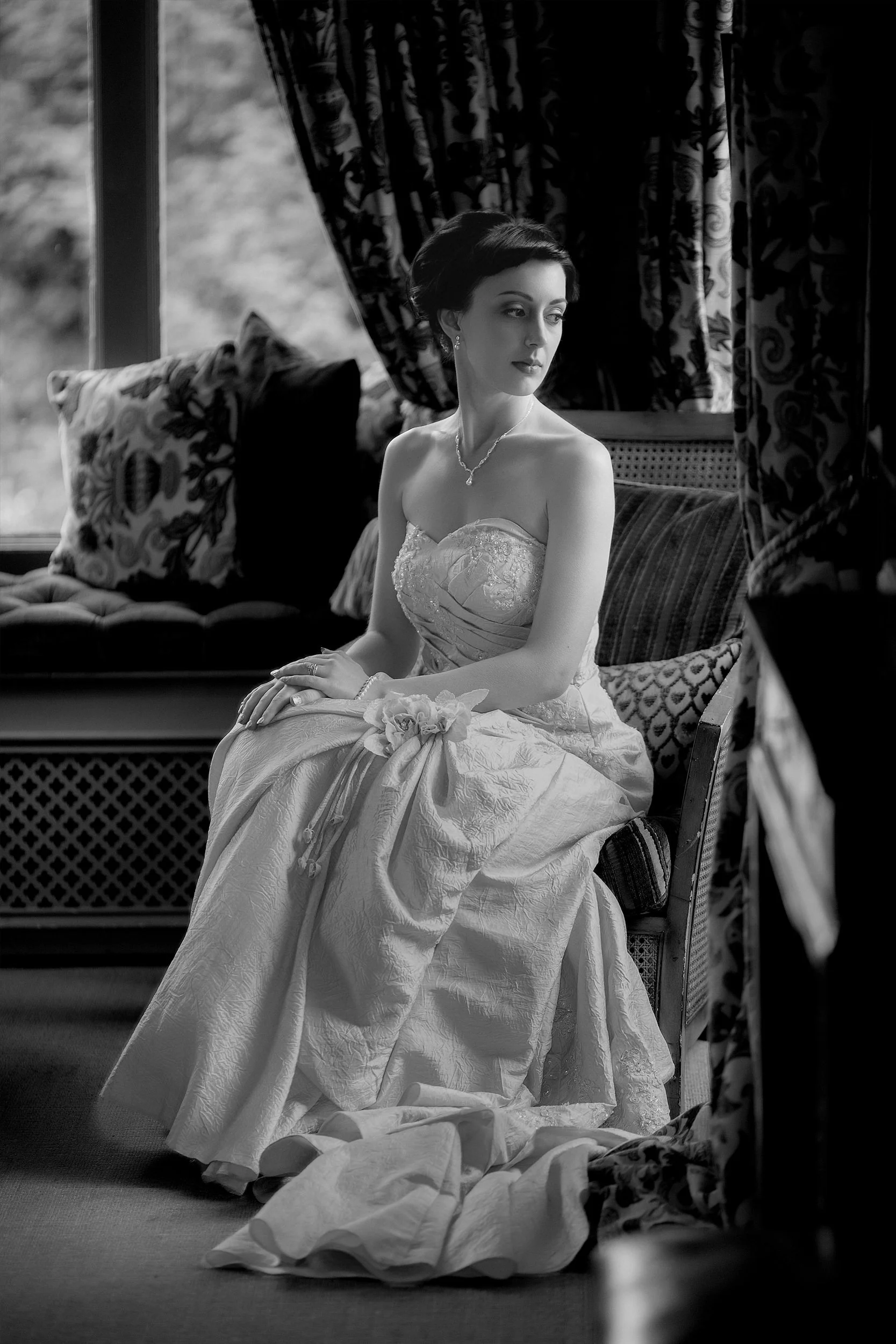 A woman in a wedding dress sitting on an ornate couch in a room with patterned curtains and pillows, looking away thoughtfully.